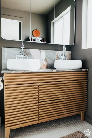 Double vanity featuring a slatted wood-finish cabinet, dual white vessel sinks, stone-surface countertop and backsplash, and wall-mounted faucets