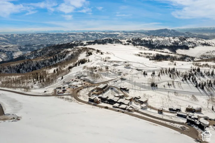 Expansive snow-covered landscape with distant mountain ranges, featuring a cluster of residential structures, a winding paved road, and scattered evergreen trees