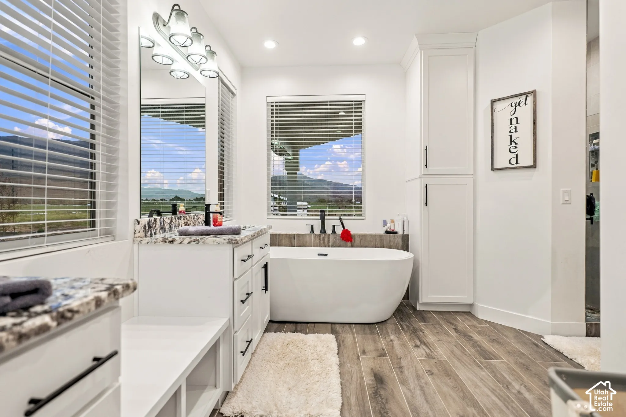 Freestanding soaking tub, wood-finish flooring, two large windows, white cabinetry with black hardware, and granite-style countertops