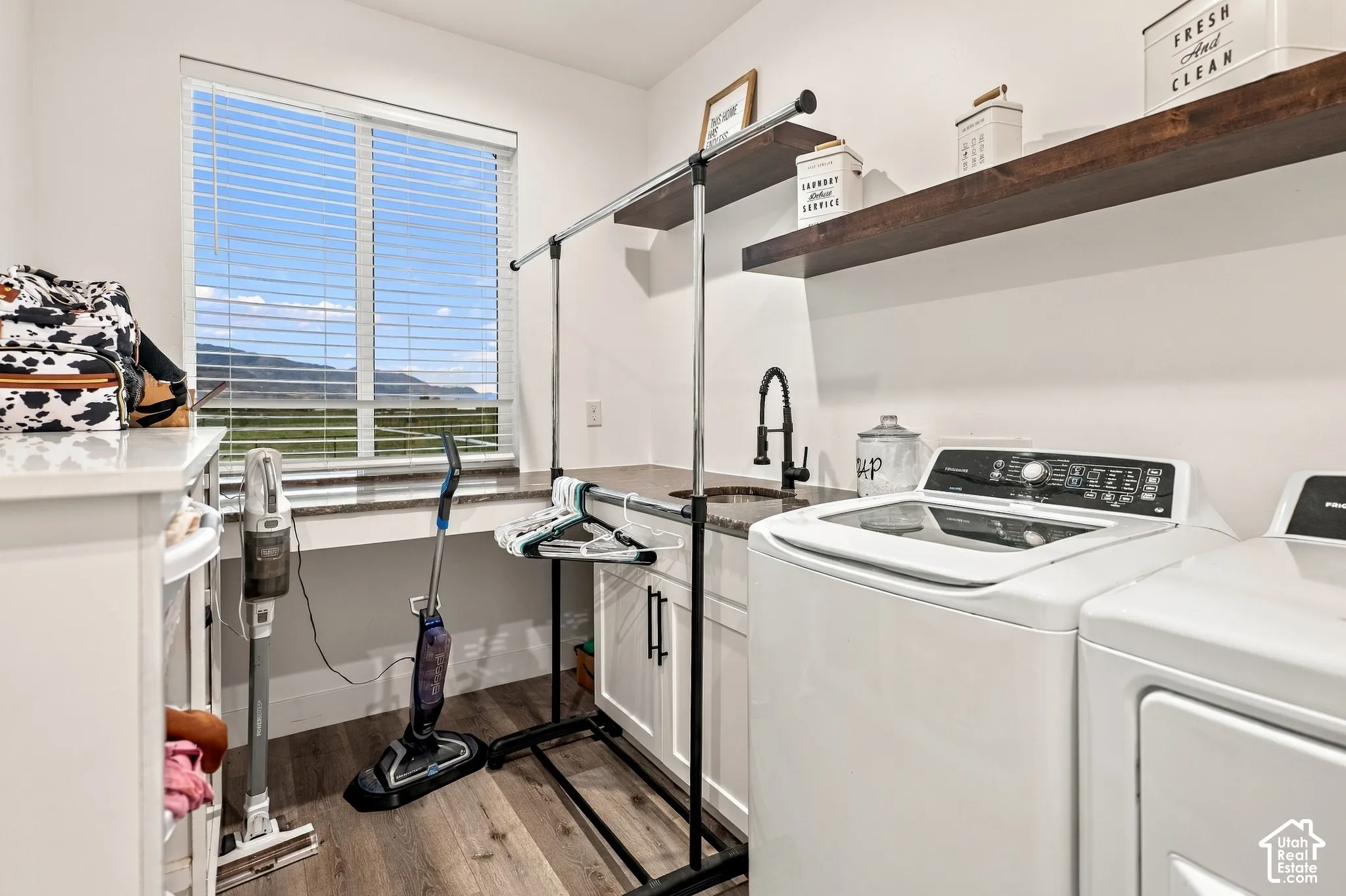 Laundry area with wood-finish flooring, white cabinetry, a built-in sink with a gooseneck faucet, and two dark wood floating shelves