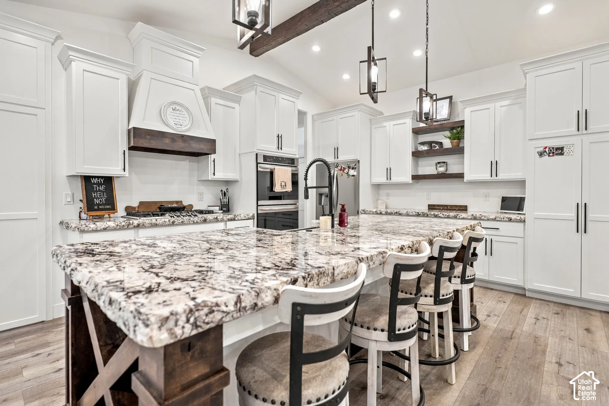 White shaker cabinetry, two-tone kitchen island with stone countertop, wood-finish flooring, built-in double ovens, and exposed ceiling beam