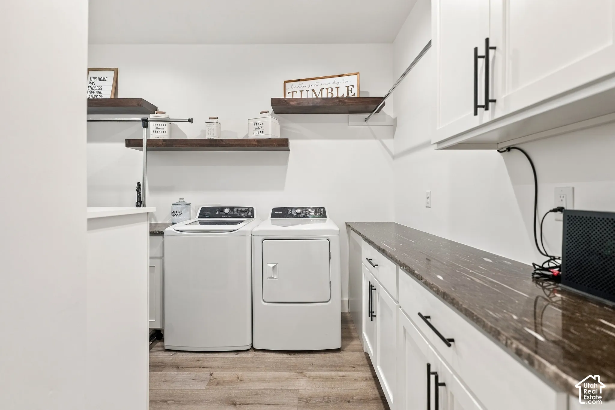 Laundry area featuring wood-finish flooring, white cabinetry with matte black hardware, a brown countertop, and floating wood shelves