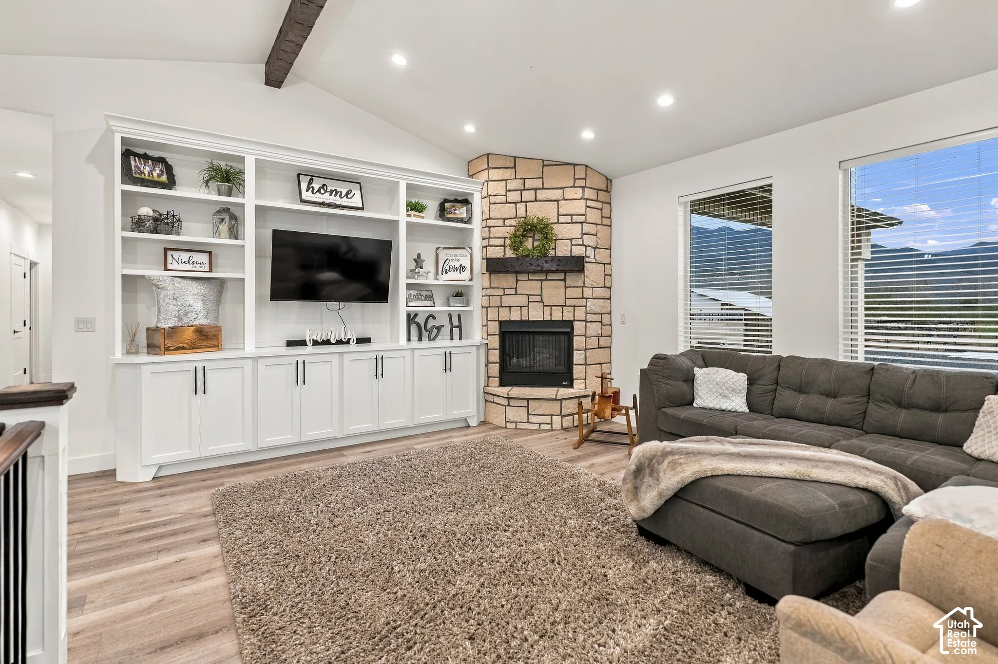 Vaulted ceiling with exposed beam, built-in entertainment center, stone fireplace, wood-finish flooring, and large windows