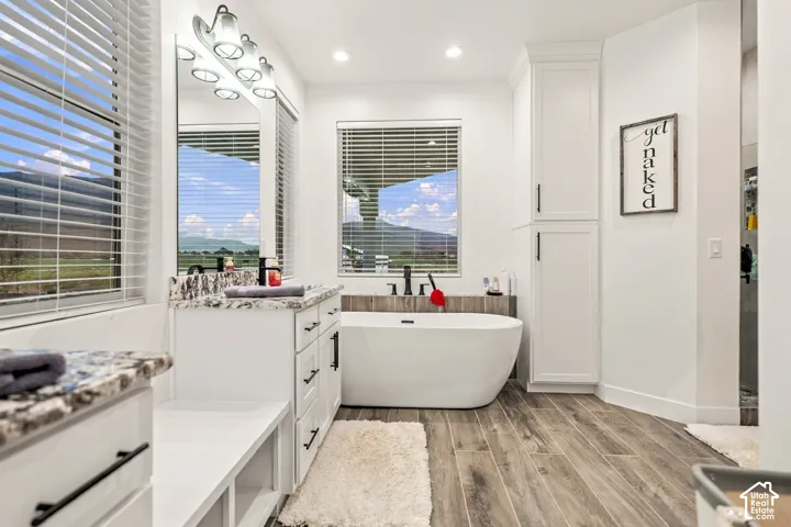 Freestanding soaking tub, wood-finish flooring, two large windows, white cabinetry with black hardware, and granite-style countertops