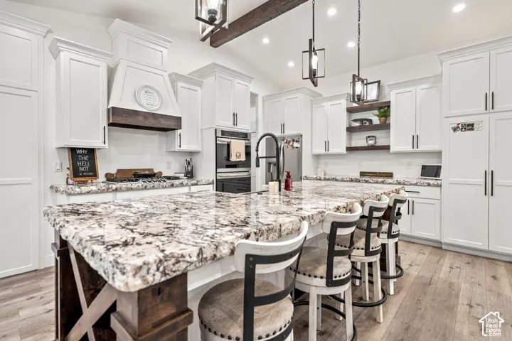 White shaker cabinetry, two-tone kitchen island with stone countertop, wood-finish flooring, built-in double ovens, and exposed ceiling beam