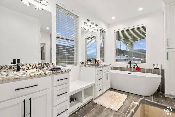 Dual vanities with stone countertops, a freestanding soaking tub, wood-look tile flooring, and white cabinetry