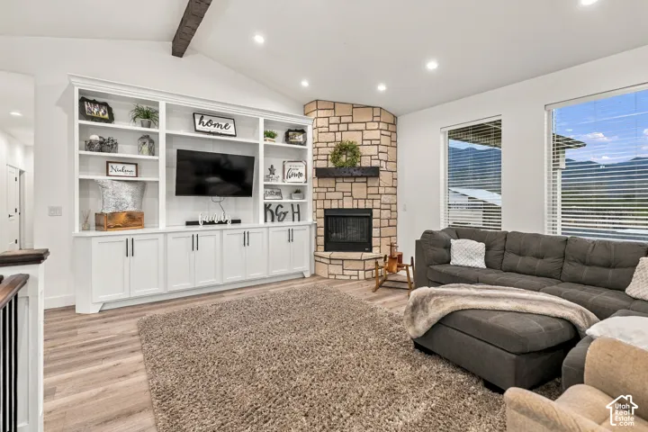Vaulted ceiling with exposed beam, built-in entertainment center, stone fireplace, wood-finish flooring, and large windows