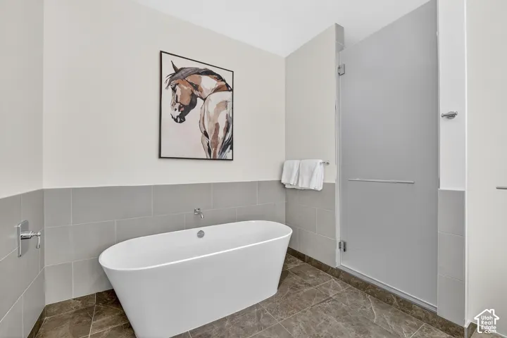 Freestanding soaking tub with wall-mounted faucet, gray tiled walls, and a glass-enclosed shower with a full-height solid panel