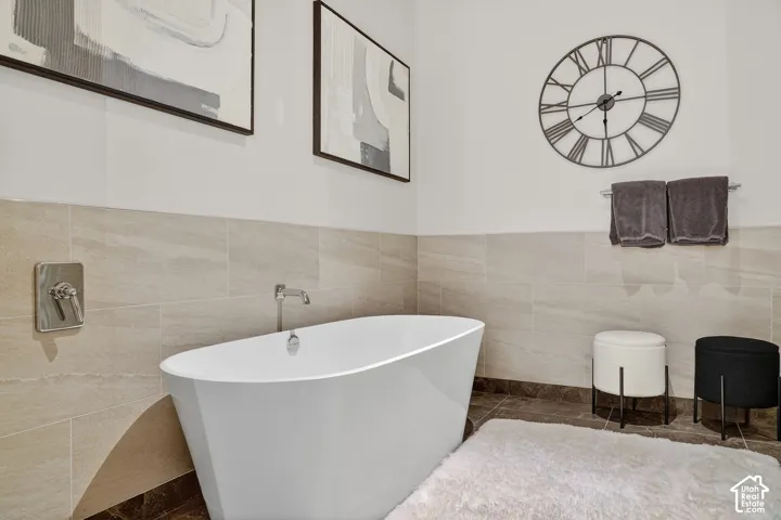 Freestanding soaking tub with wall-mounted faucet, neutral-toned tiled walls, and coordinating floor tiles