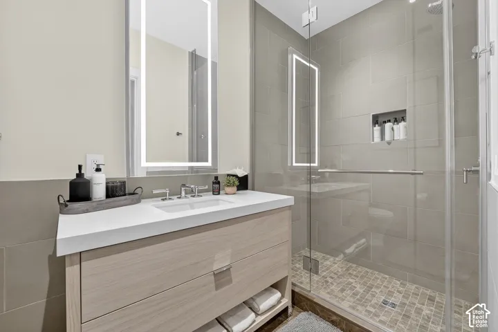Vanity with integrated sink and chrome faucet, backlit mirror, glass-enclosed shower, shower niche, and mosaic tile shower floor