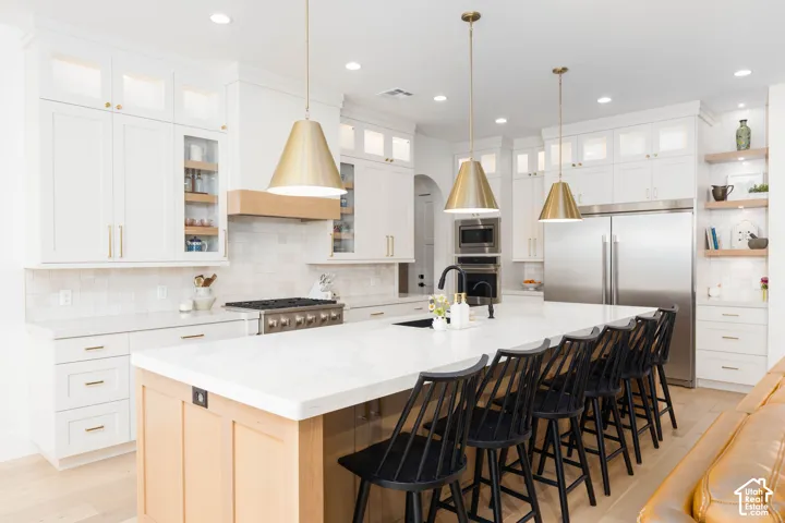 White cabinetry with glass-front accents, stainless steel appliances, a center island with seating, a tile backsplash, and light wood-finish flooring