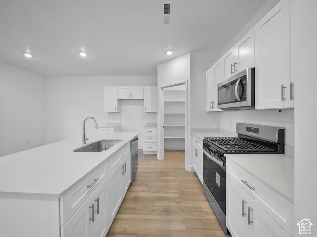 Kitchen featuring white cabinetry, stainless steel appliances, a center island with an integrated sink, light-toned countertops, and wood-finish flooring