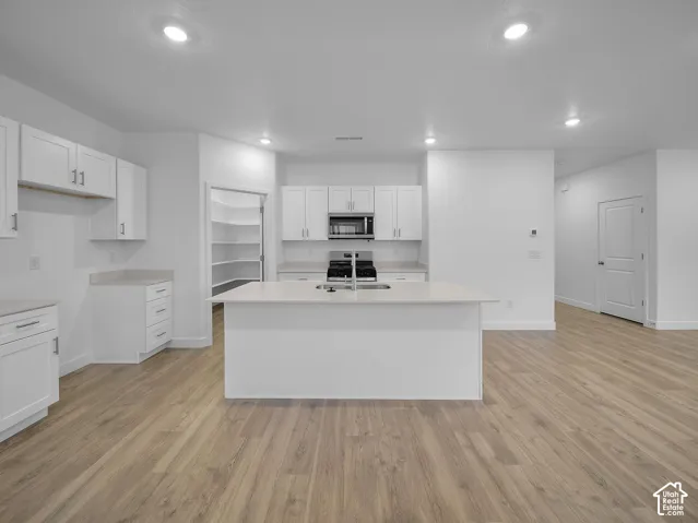 Kitchen featuring white cabinetry, light-toned countertops, a central island with an integrated sink, a built-in microwave, and wood-finish flooring