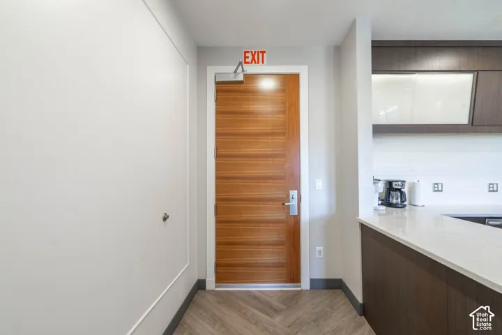 Wood-finish door with brushed nickel hardware, herringbone wood-finish flooring, white tile backsplash, light-toned countertop, and dark wood-finish cabinetry