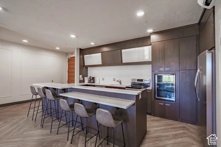 Modern kitchen featuring dark wood-finish cabinetry, stainless steel appliances, a built-in beverage refrigerator, and an expansive breakfast bar with light-toned countertops