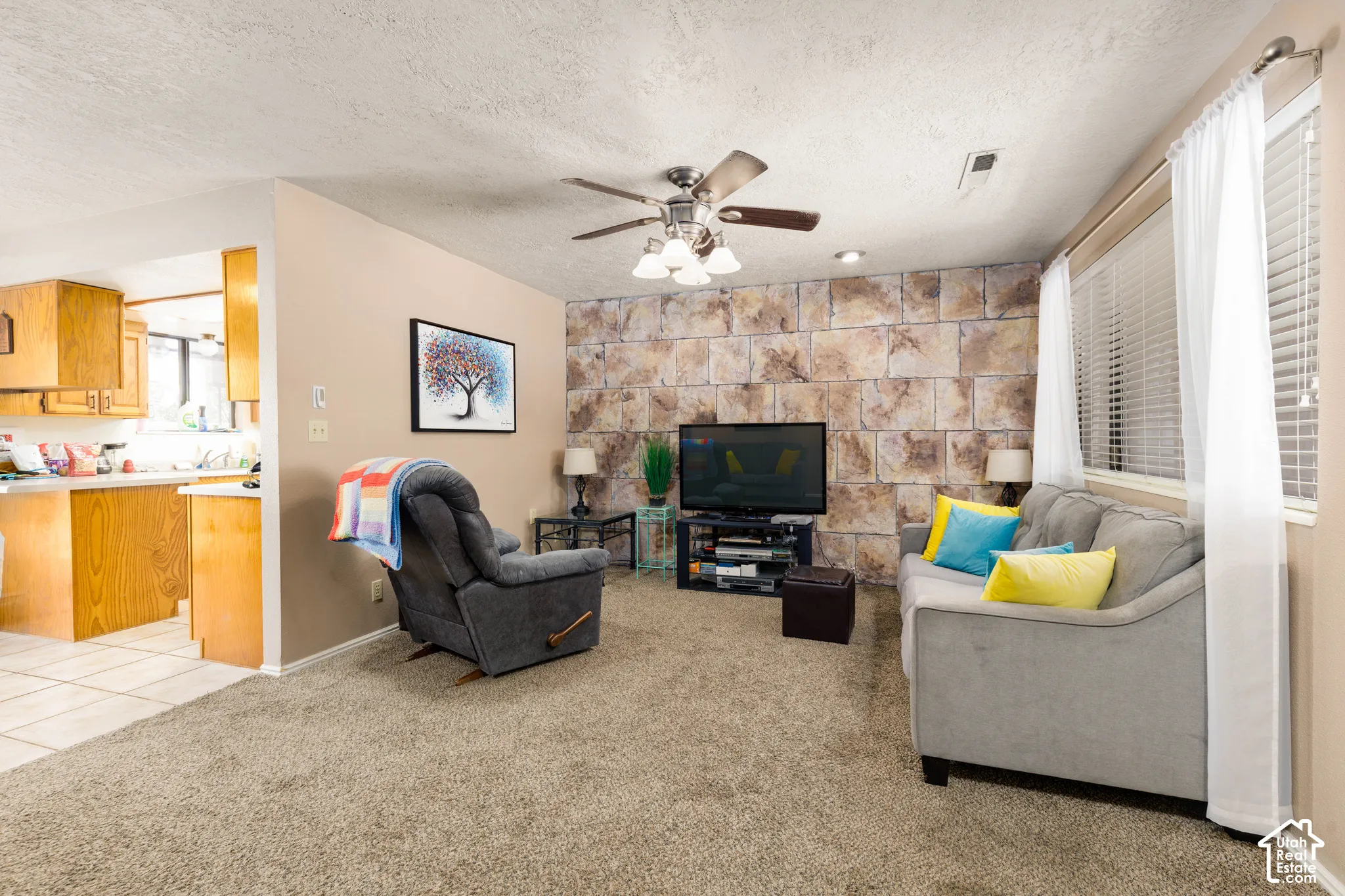 Textured accent wall, ceiling fan with integrated lighting, window with blinds and sheer curtains, carpeted flooring, and a kitchen visible with wood cabinetry