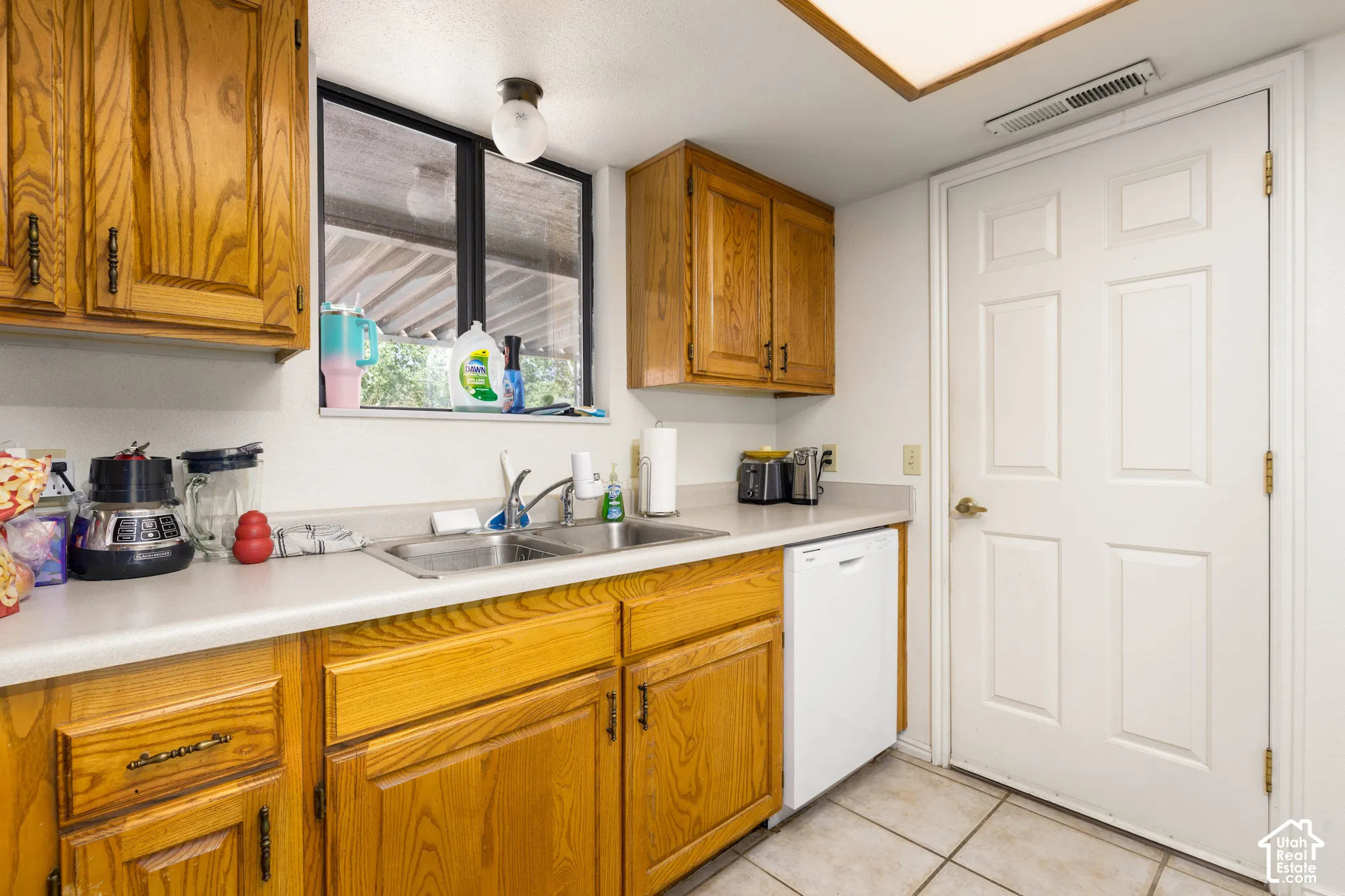 Wood cabinetry, laminate countertops, a stainless steel double basin sink, and a white dishwasher