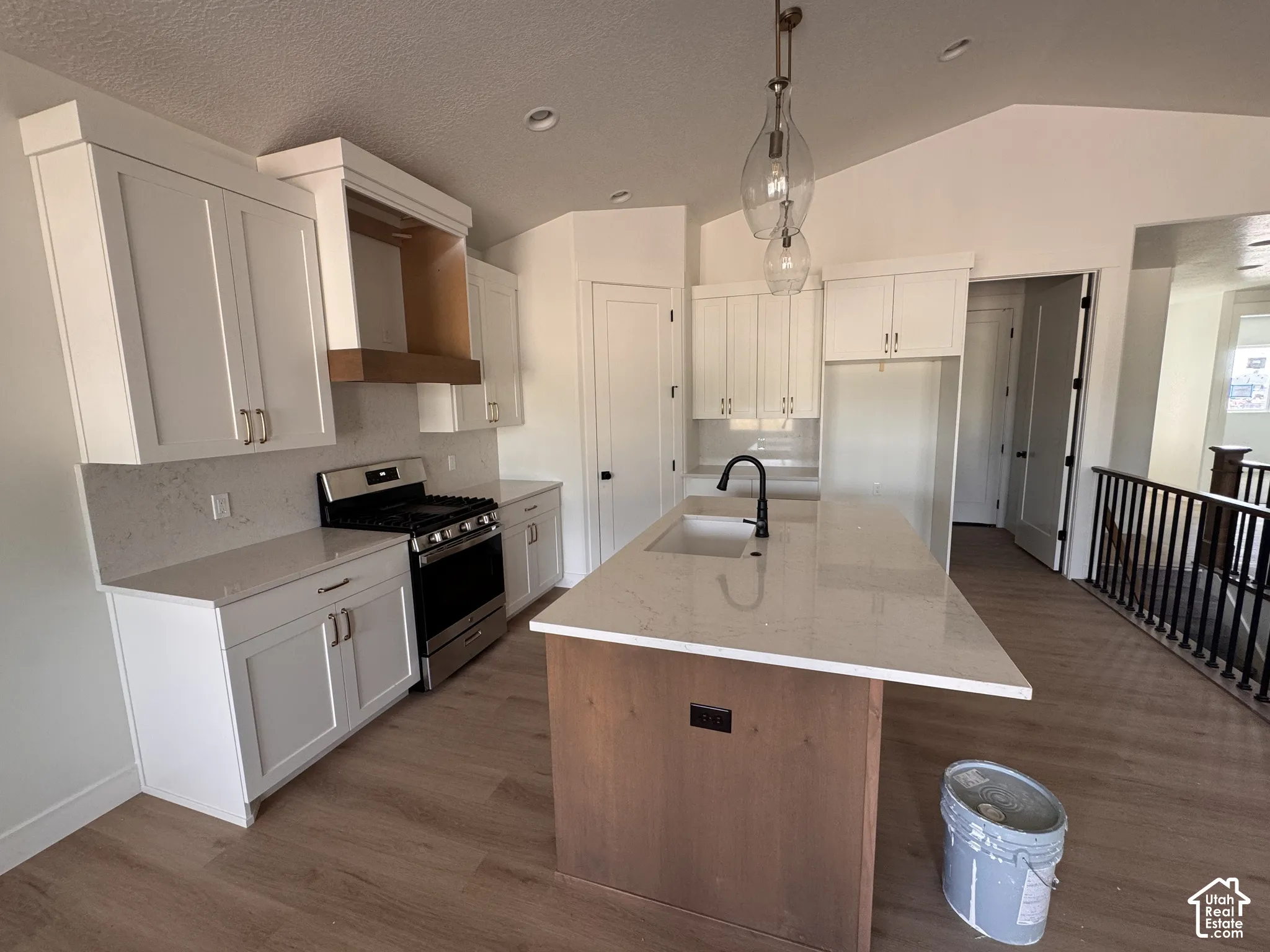 Kitchen featuring white shaker cabinetry, stainless steel gas range, light-toned countertops, a central island with an integrated sink and matte black faucet, and wood-finish flooring