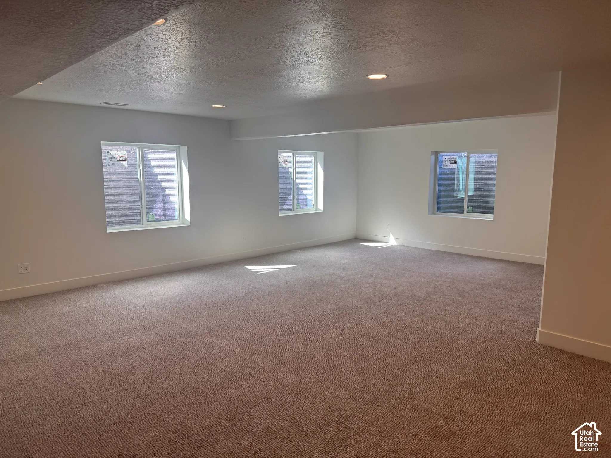 Spacious open floor plan featuring neutral carpeting, recessed ceiling lighting, and abundant natural light from three large windows with plantation shutters