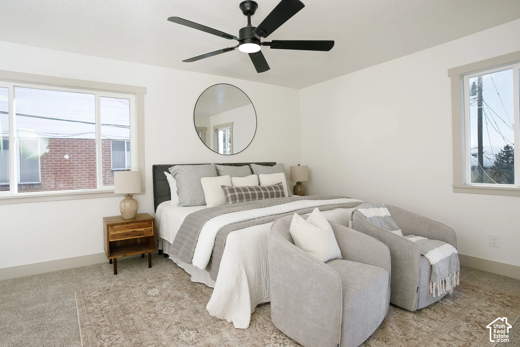 White walls with light gray trim, two large windows, black ceiling fan with integrated lighting, and neutral carpeting