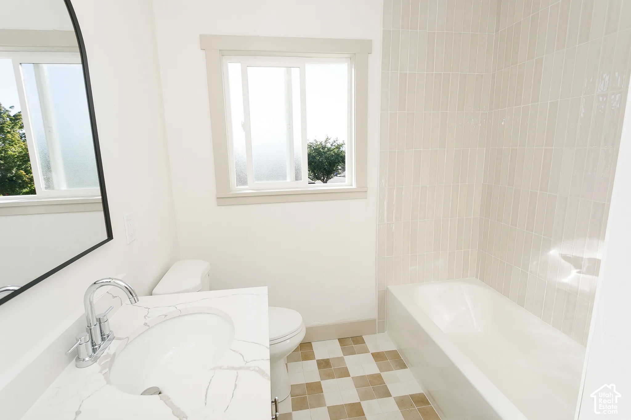 White vanity with integrated sink and chrome faucet, checkered tile flooring, white bathtub with vertical tile surround, and an arched mirror