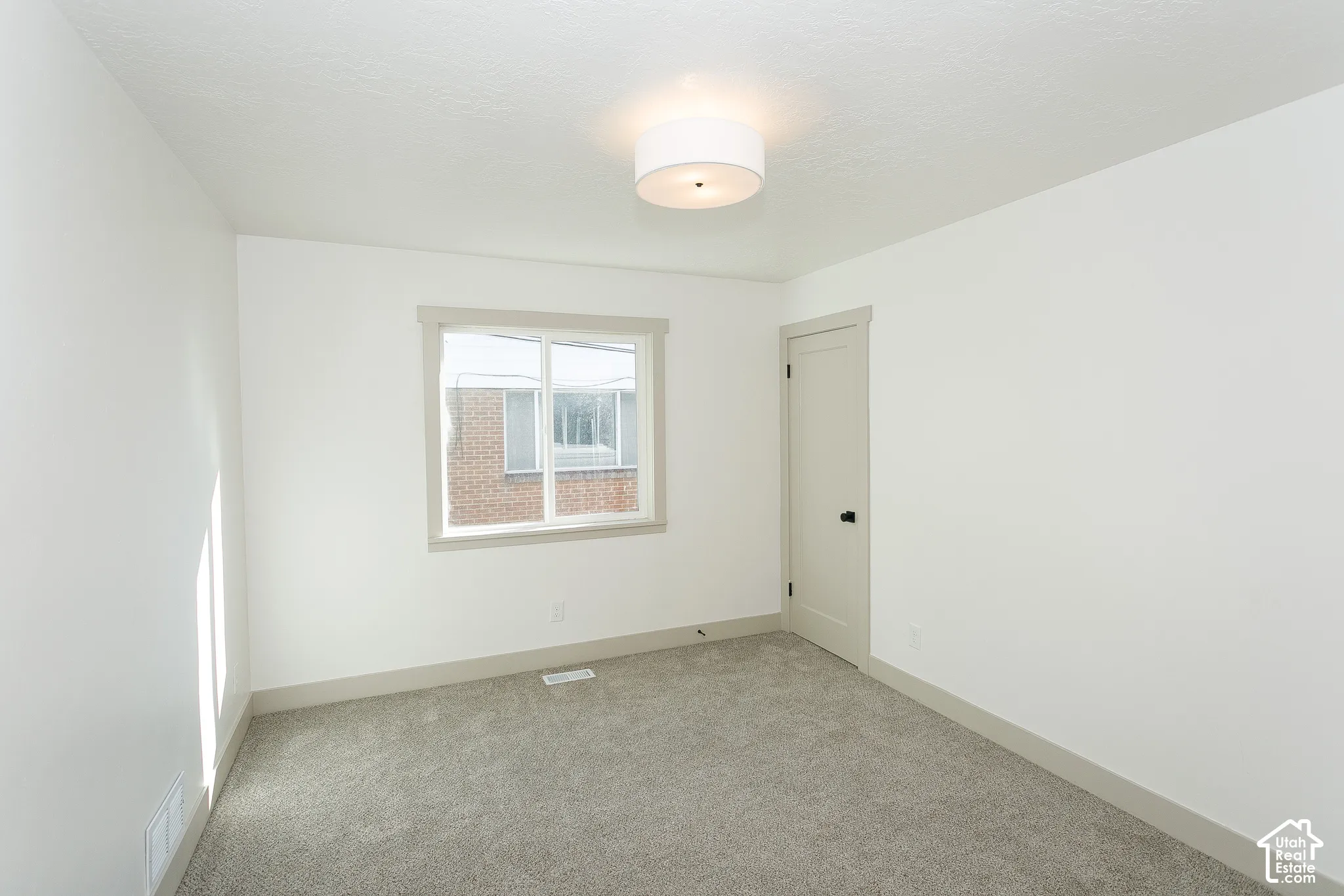 White walls, grey carpet, window with light-colored trim, light-colored door with black hardware, and a semi-flush ceiling light fixture