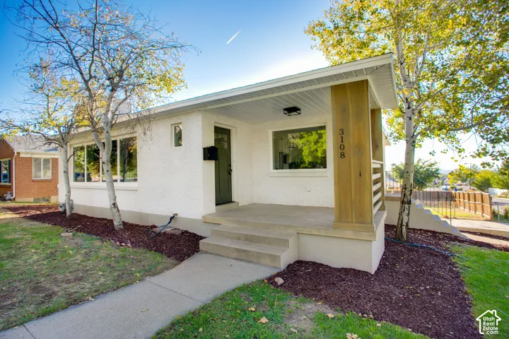 White stucco exterior, covered front porch with wood-clad columns, concrete steps, and a manicured landscape featuring young trees and ground cover
