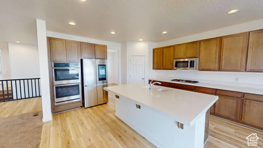 Expansive kitchen island with a built-in sink, stainless steel appliances, wood-finish cabinetry, light wood-finish flooring, and recessed lighting