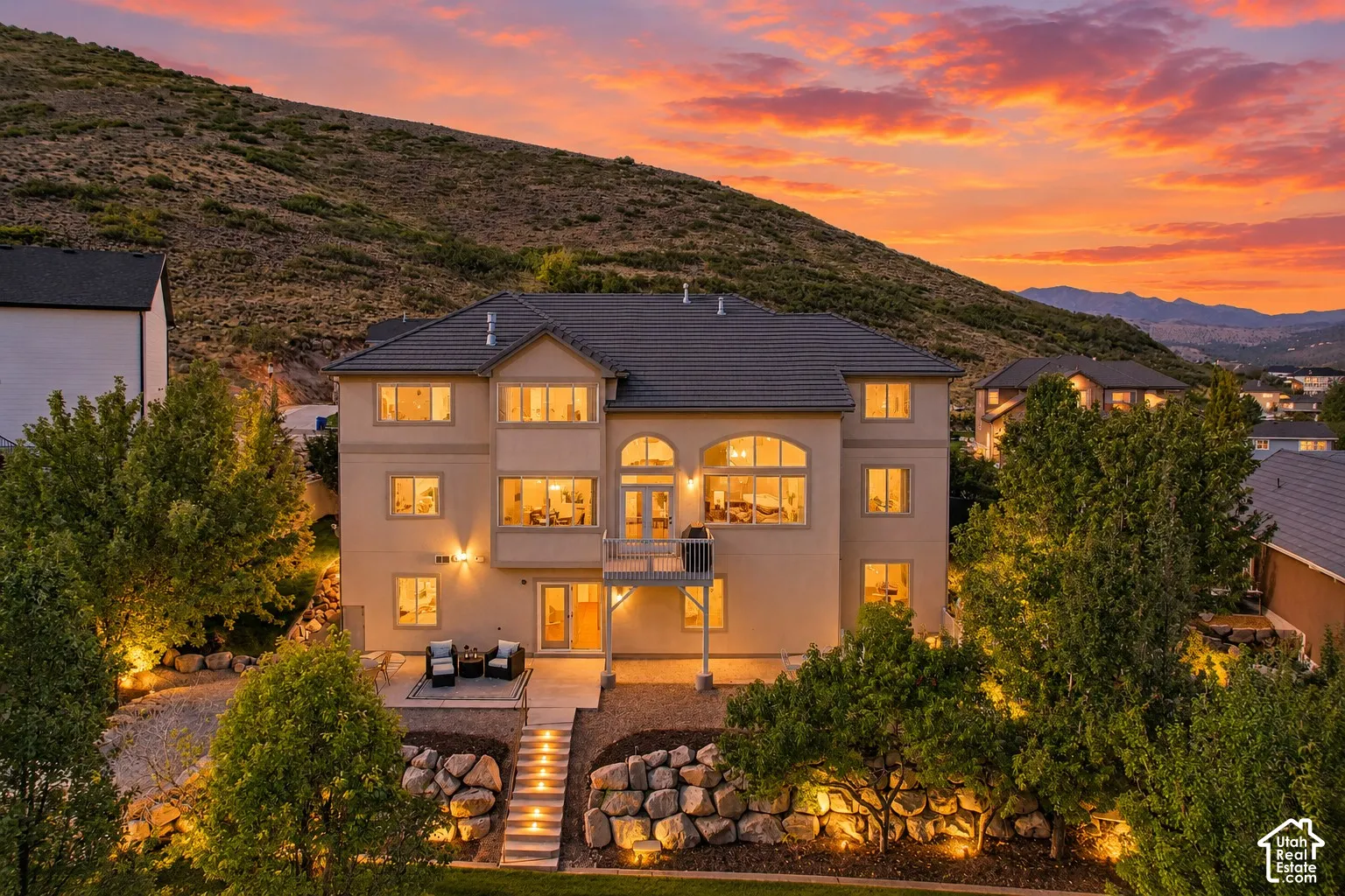 Stucco exterior with multiple windows, tiered landscaping, a paved patio, and a second-story balcony