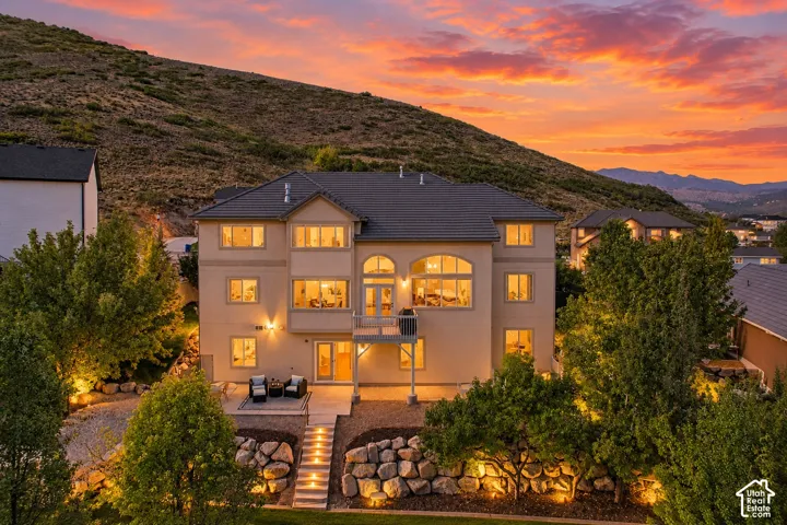 Stucco exterior with multiple windows, tiered landscaping, a paved patio, and a second-story balcony