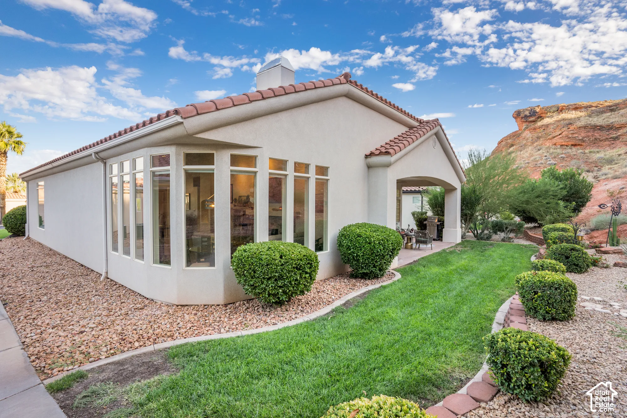 Stucco exterior with a tile roof, featuring a bay window, a covered patio, and a landscaped backyard with a manicured lawn and decorative plantings
