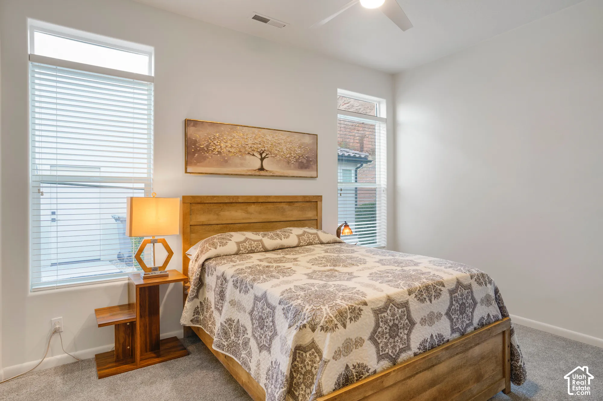 Neutral wall color, light wood-finish bed frame, tall windows with blinds, ceiling fan, and carpeted flooring