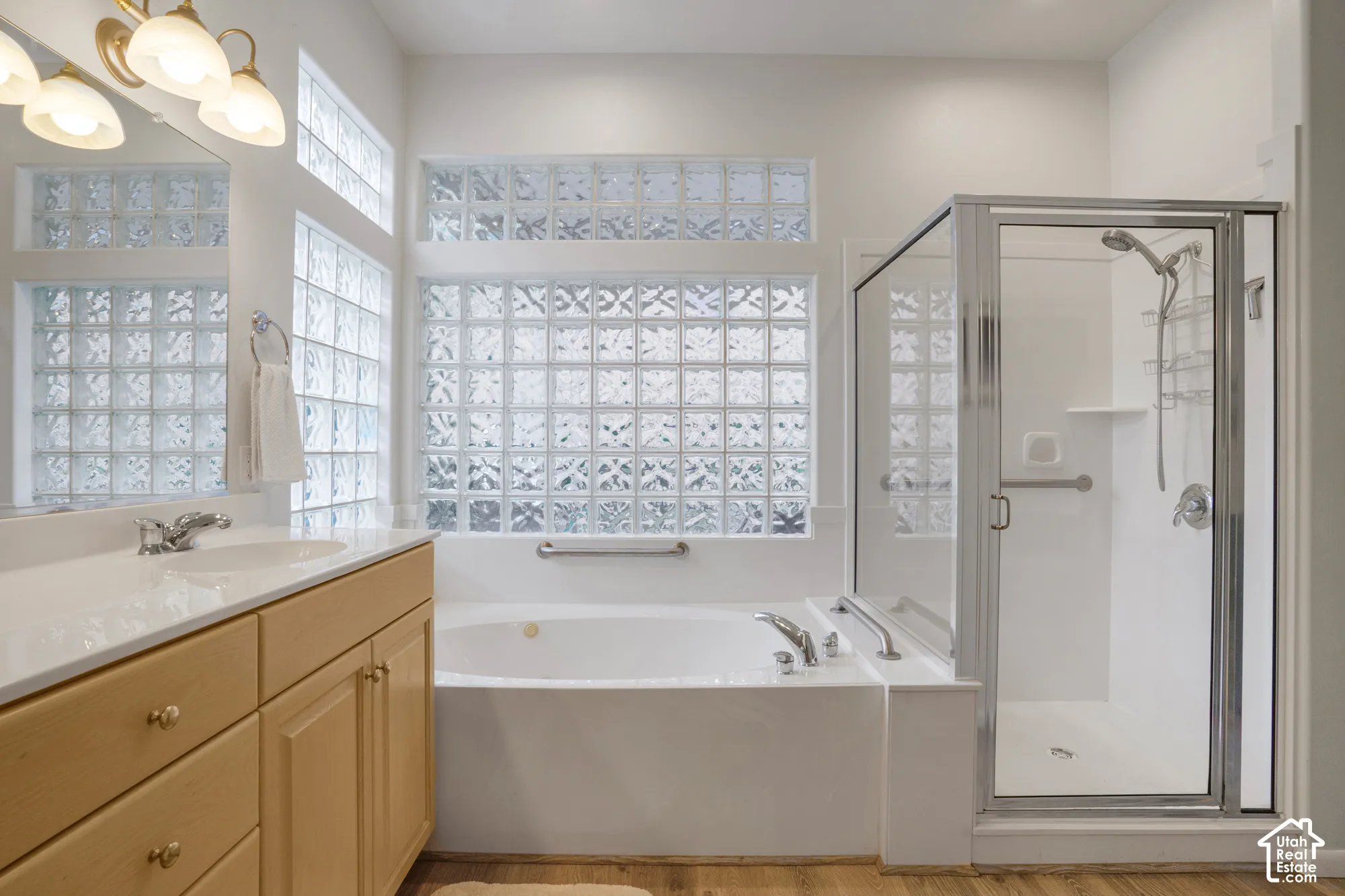 Bathroom featuring a double vanity with light wood cabinetry, a built-in soaking tub, a separate glass-enclosed shower, and decorative glass block windows