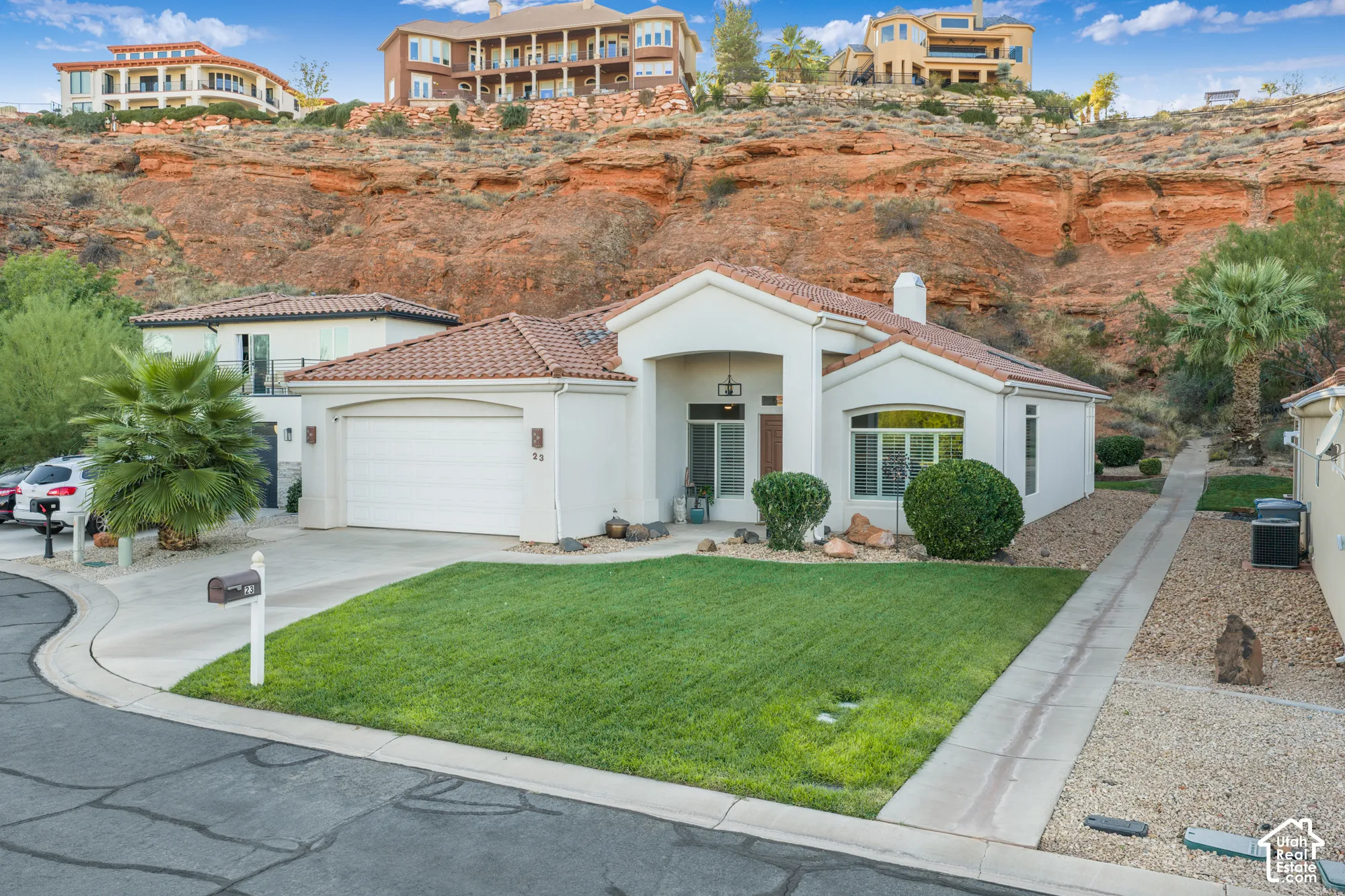 Contemporary single-story residence featuring a clean white stucco exterior with a brown tile roof, positioned on a sloped lot with dramatic red-rock backdrop