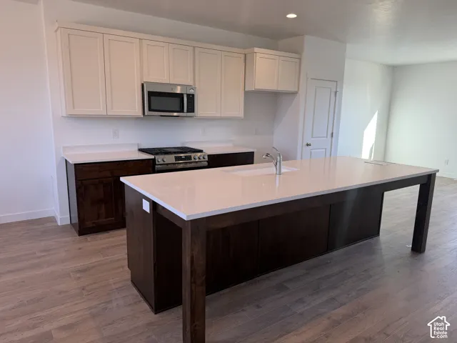 Kitchen featuring wood-finish flooring, two-tone cabinetry, stainless steel appliances, a center island with an integrated sink, and light-toned countertops