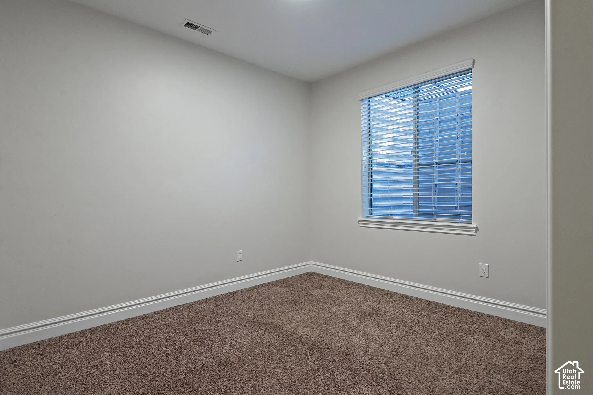 Bright corner bedroom featuring neutral wall finishes, carpet flooring, and a large window with plantation shutters allowing natural light throughout the space