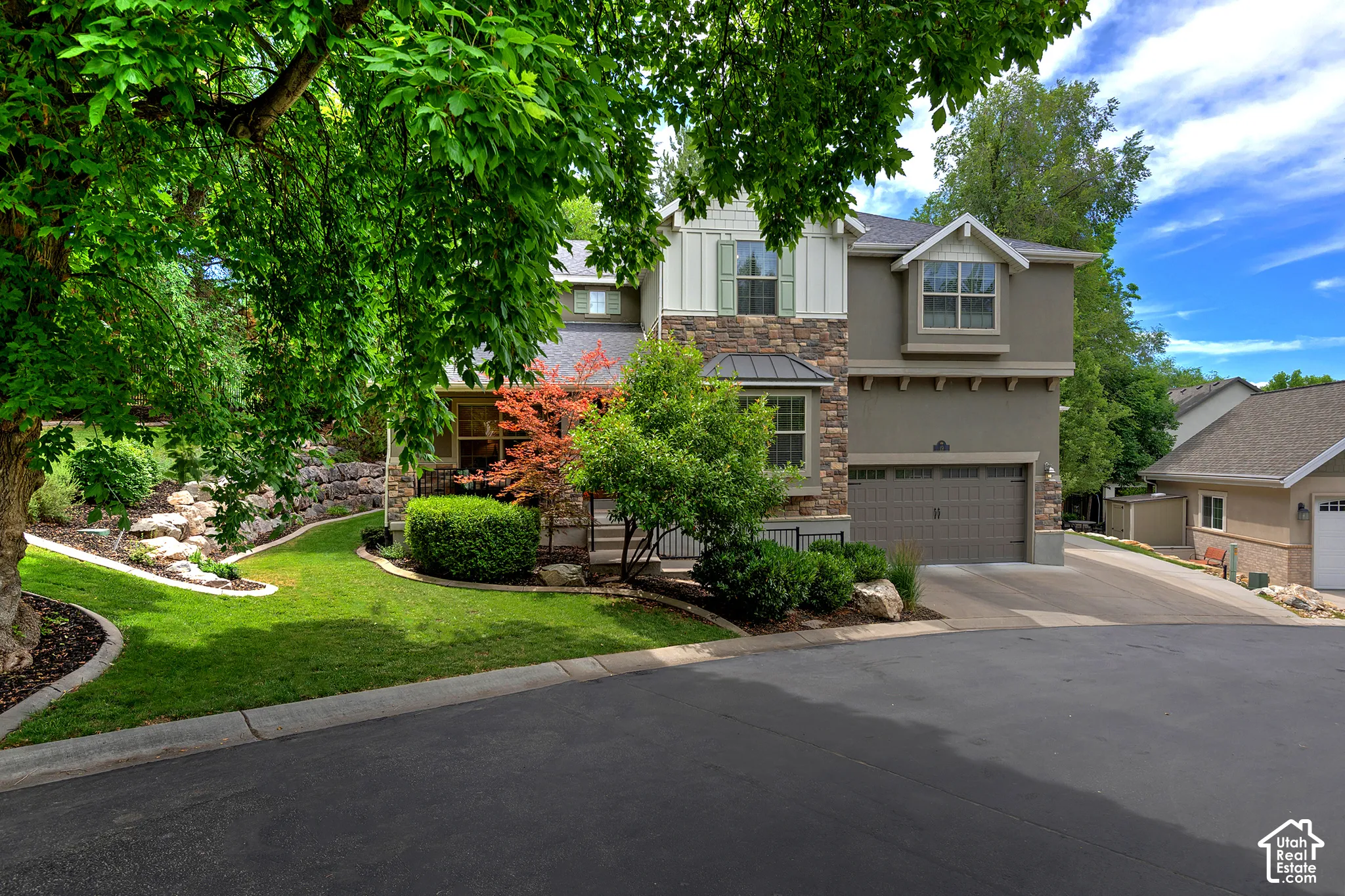Two-story residence with taupe exterior and stone accents, featuring a three-car garage, mature landscaping with manicured lawn, and established shade trees framing the property