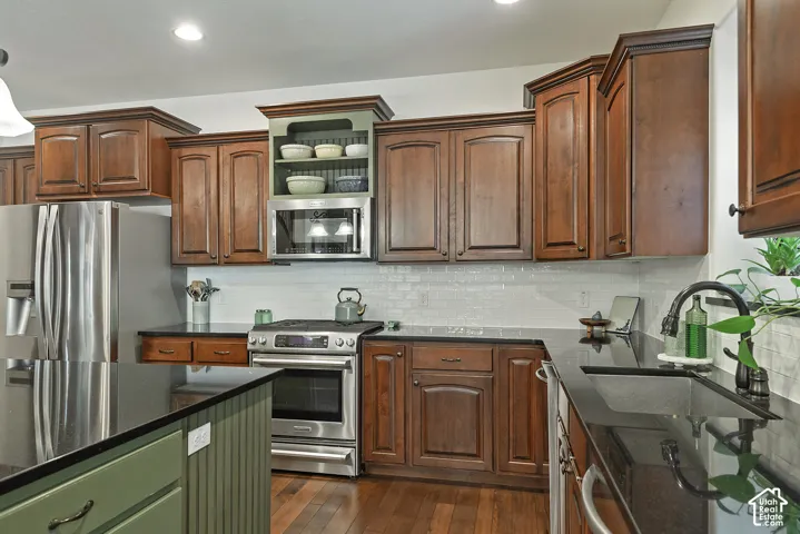 Kitchen featuring rich wood cabinetry, dark countertops, a subway tile backsplash, stainless steel appliances, and wood-finish flooring
