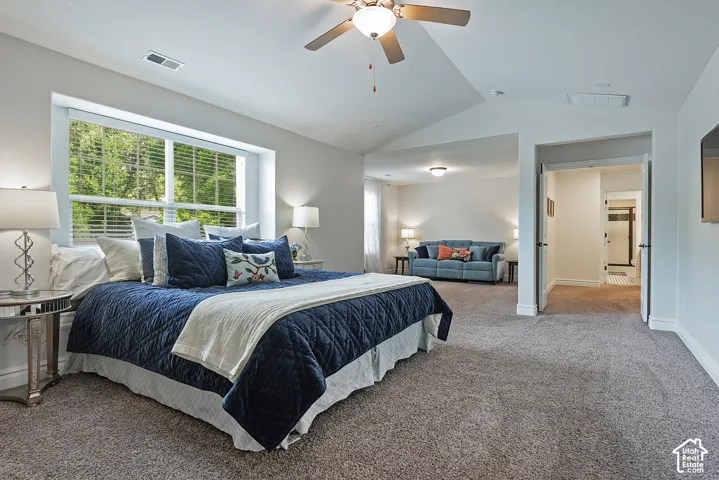 Vaulted ceiling, ceiling fan with integrated lighting, large window with blinds, carpeted flooring, and an open doorway revealing an additional room
