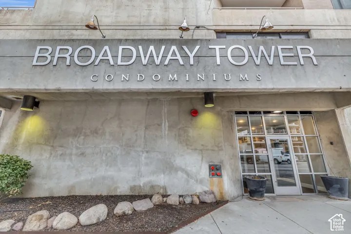 Concrete facade with "Broadway Tower Condominiums" signage, ground-level entrance with glass doors, and exterior wall-mounted lighting fixtures