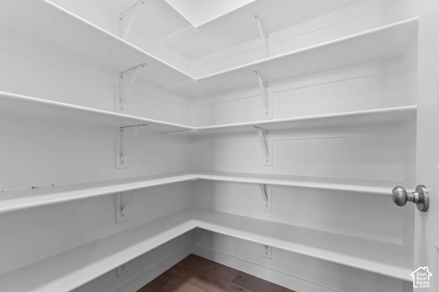 Corner pantry with multiple white shelves, wood-finish flooring, and a white door with a brushed nickel knob