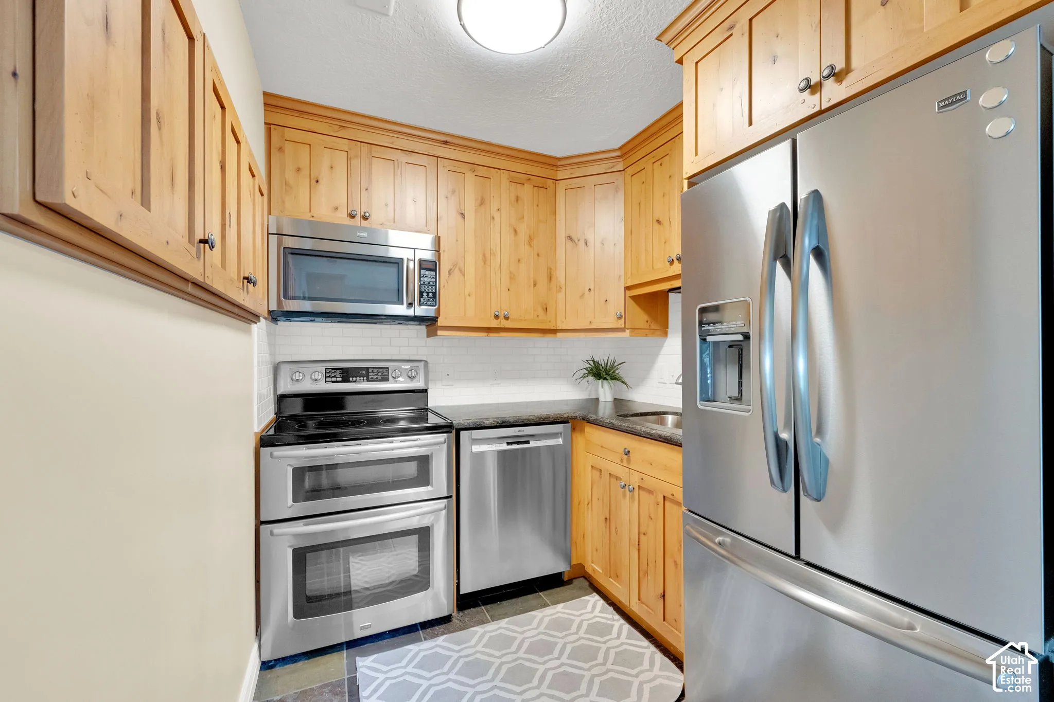 Wood cabinetry with black hardware, stainless steel appliances, white subway tile backsplash, and dark countertops