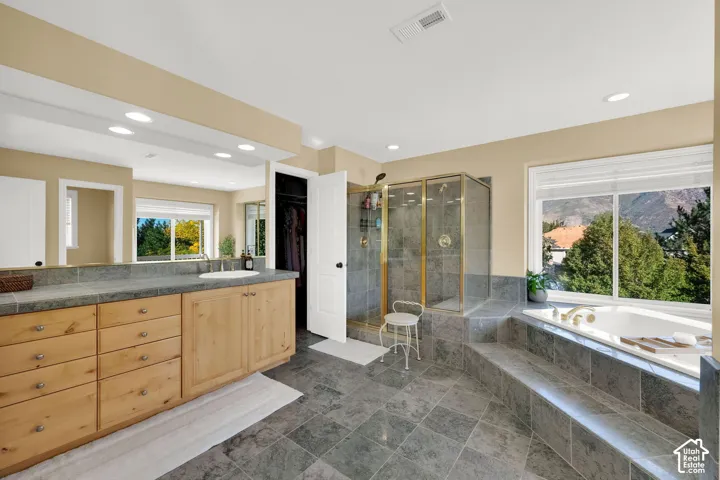 Spacious bathroom featuring natural wood vanity cabinetry, stone countertop, integrated soaking tub, and a glass-enclosed shower