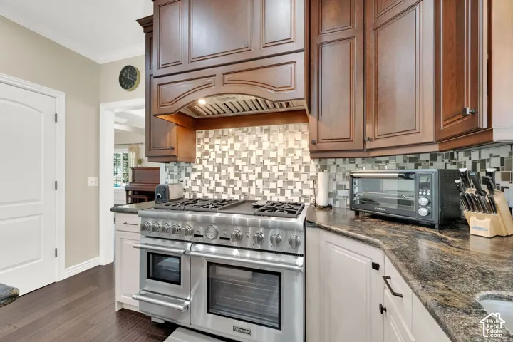 Stainless steel range with double ovens, mosaic tile backsplash, dark wood cabinetry, white lower cabinetry, and dark granite countertops