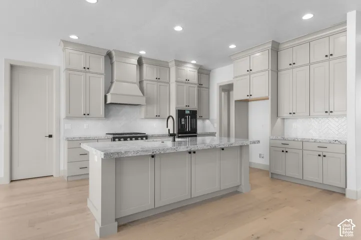 Spacious kitchen featuring an expansive center island, light wood-finish flooring, recessed lighting, white herringbone tile backsplash, and light gray cabinetry with dark hardware