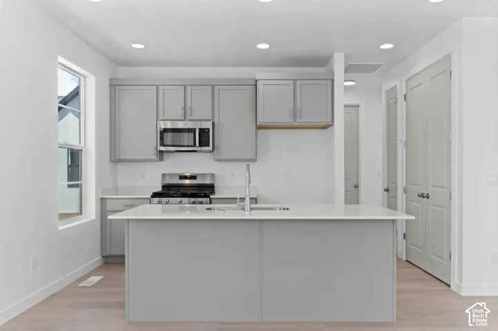 Kitchen featuring a central island with an integrated sink, light grey cabinetry, stainless steel appliances, and light-toned wood-finish flooring