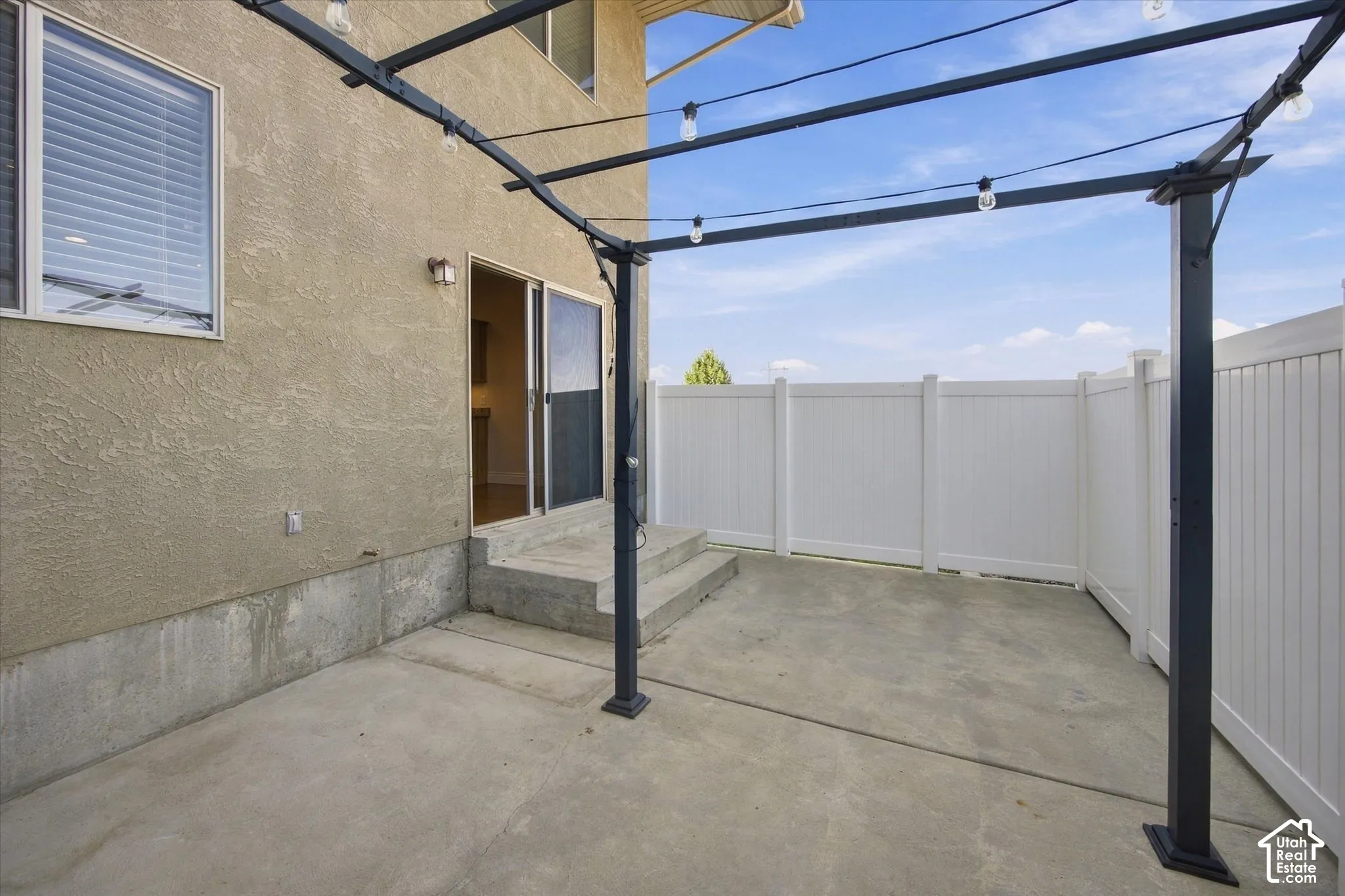 Concrete patio with a dark metal pergola, string lighting, and white privacy fencing