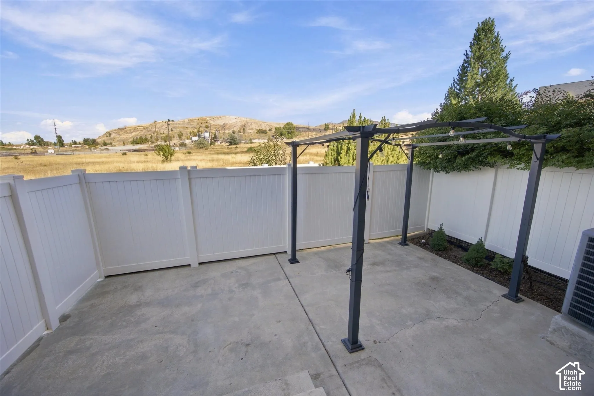 Spacious rooftop deck featuring a concrete surface, modern black metal pergola with climbing vines, white privacy fencing, and panoramic hillside backdrop with mature landscaping