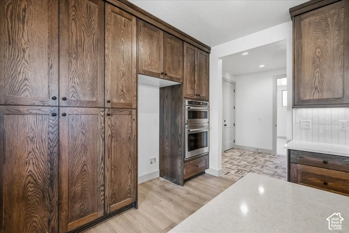 Dark wood-finish cabinetry, stainless steel double wall ovens, white subway tile backsplash, light wood-finish flooring, and a light-colored countertop