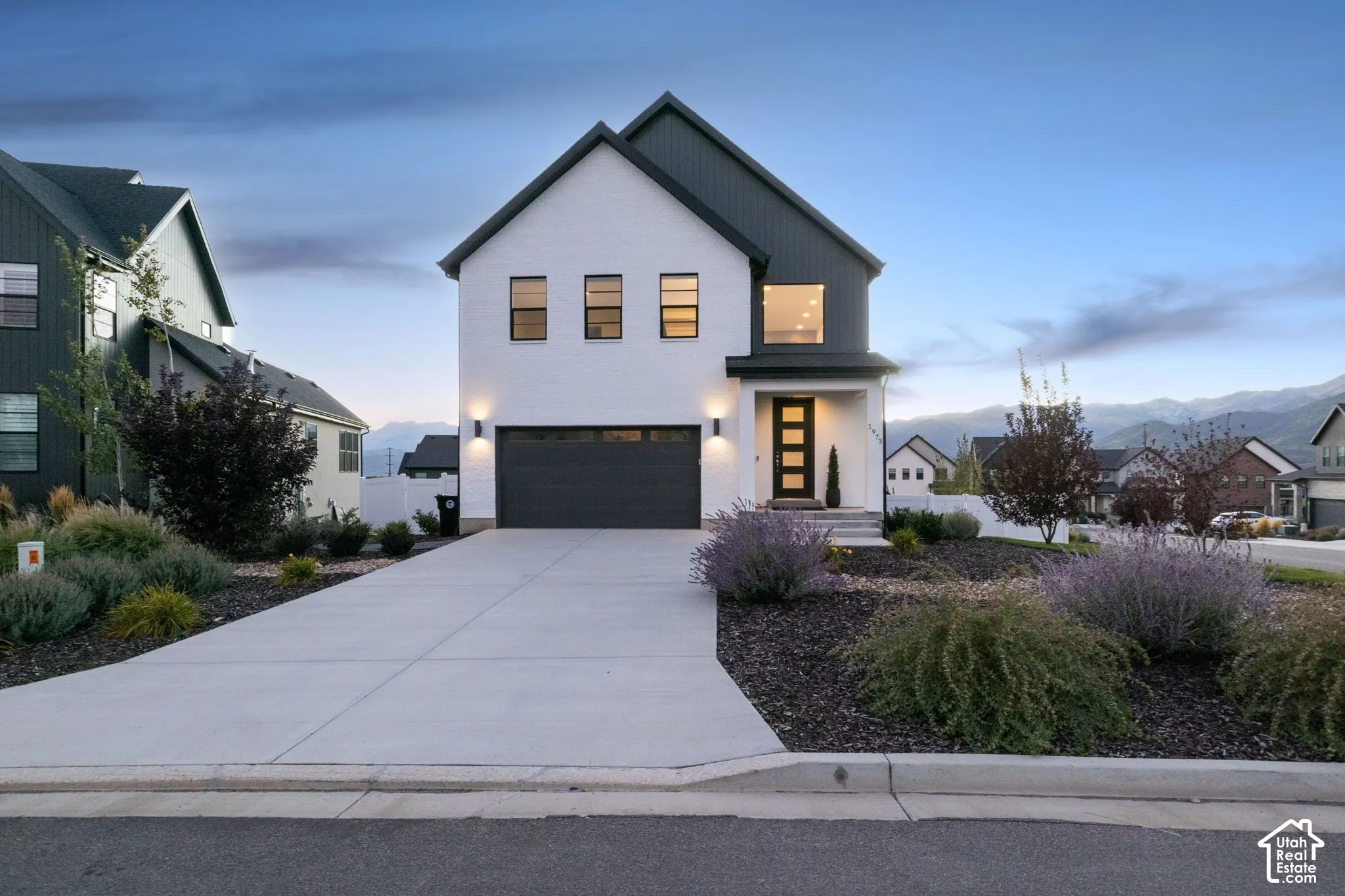 White brick exterior with dark trim accents, concrete driveway, attached garage, and landscaped front yard with varied plantings